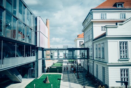 Offizielles Pressefoto des Campus der Universität für Weiterbildung Krems. Links ein Neubau aus Glas, rechts ein historisches Gebäude. Auf Höhe des ersten Stockwerks sind die beiden Gebäude durch einen Gang aus Glas verbunden. Einge Personen sind am Campus unterwegs.