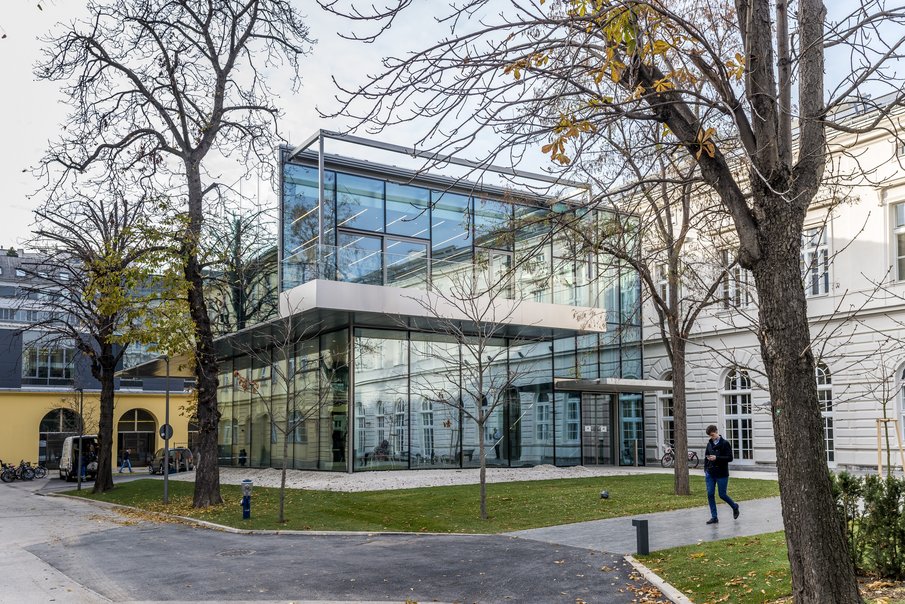 Pressefoto der mdw-Bibliothek: Ein moderner Bau mit Glasfasade, im Innenhof der Universität, umgeben von drei alten Bäumen